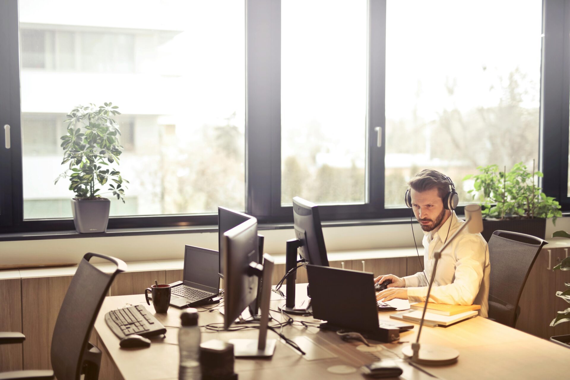 A man with headphones working on his desktop in a workplace with other desktops.